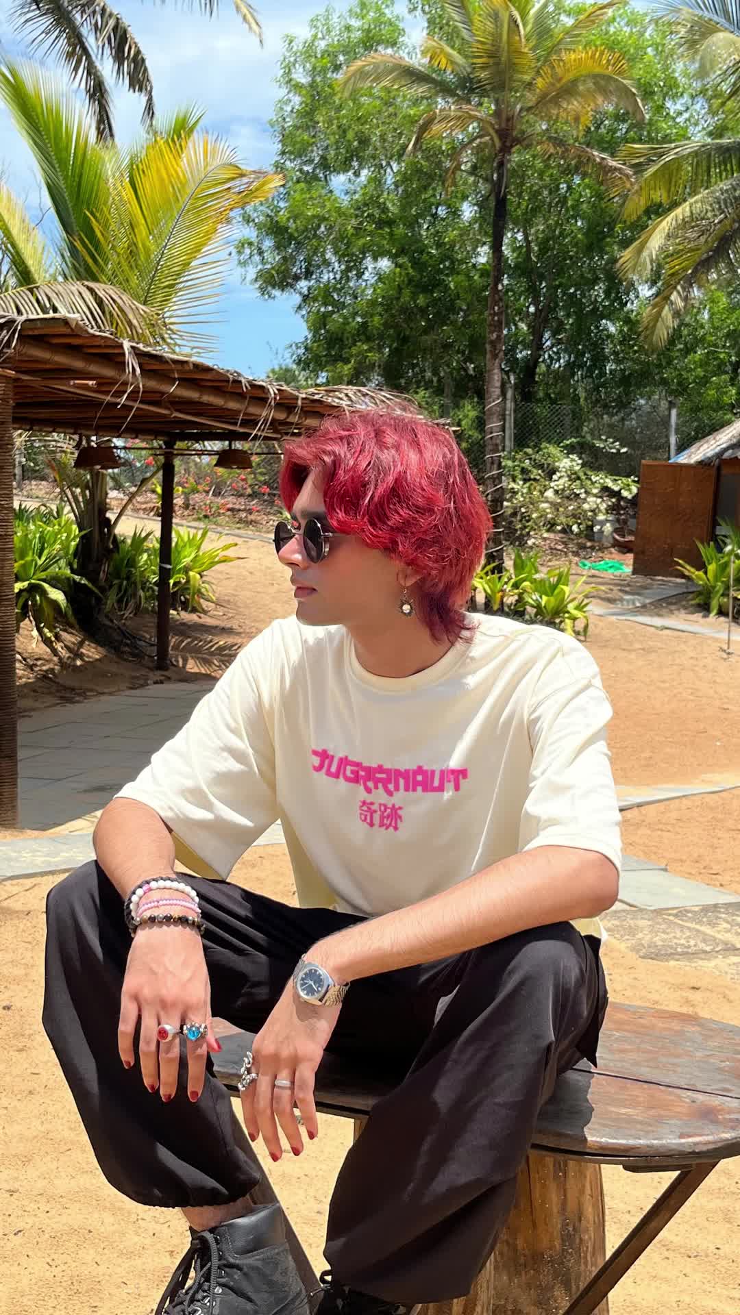 a boy with red hair sitting on a table in the middle of some forest wearing a beige tshirt and a baggy jeans