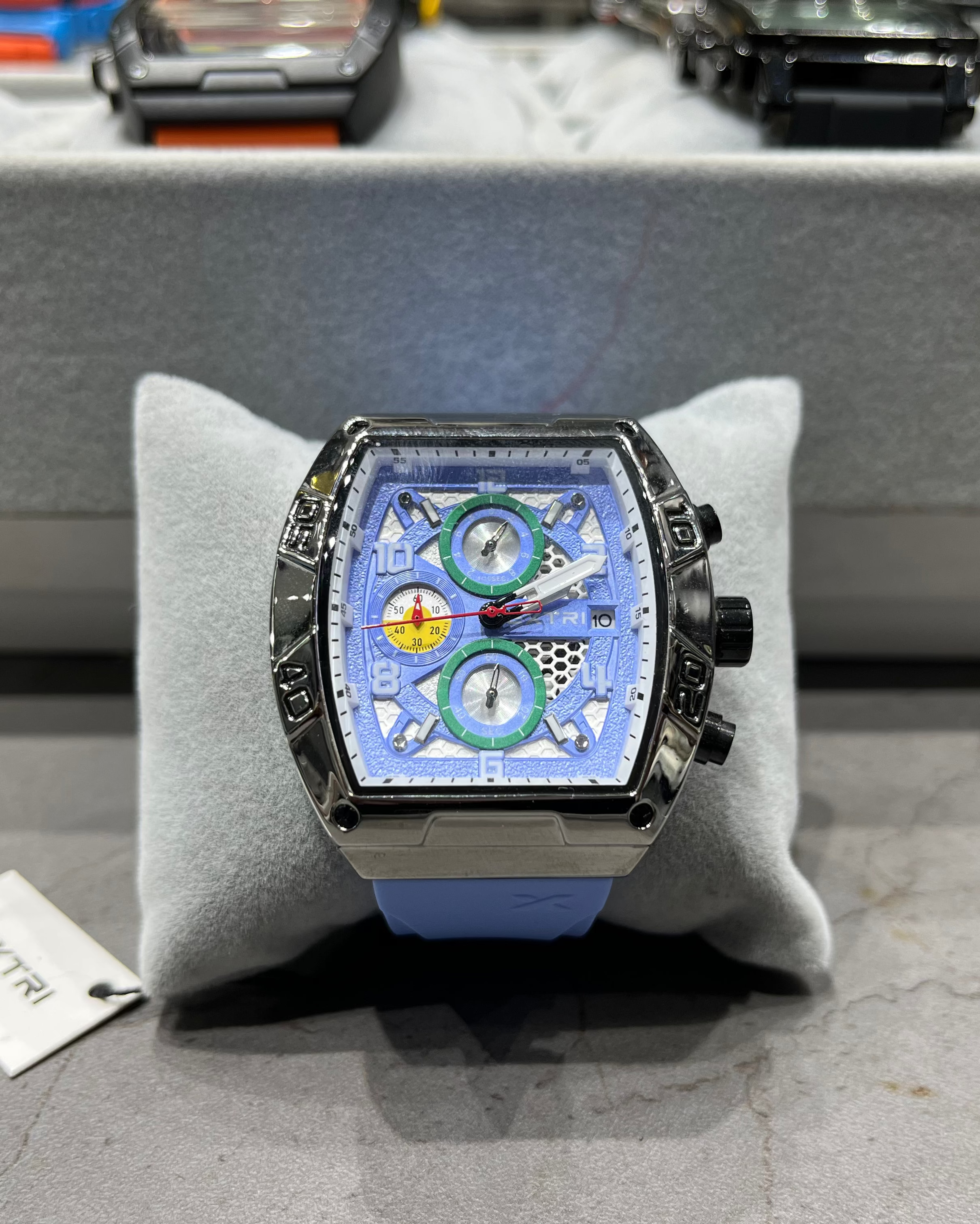 Luxury watch with a blue face on a gray cushion, with a blurred background of other watches.