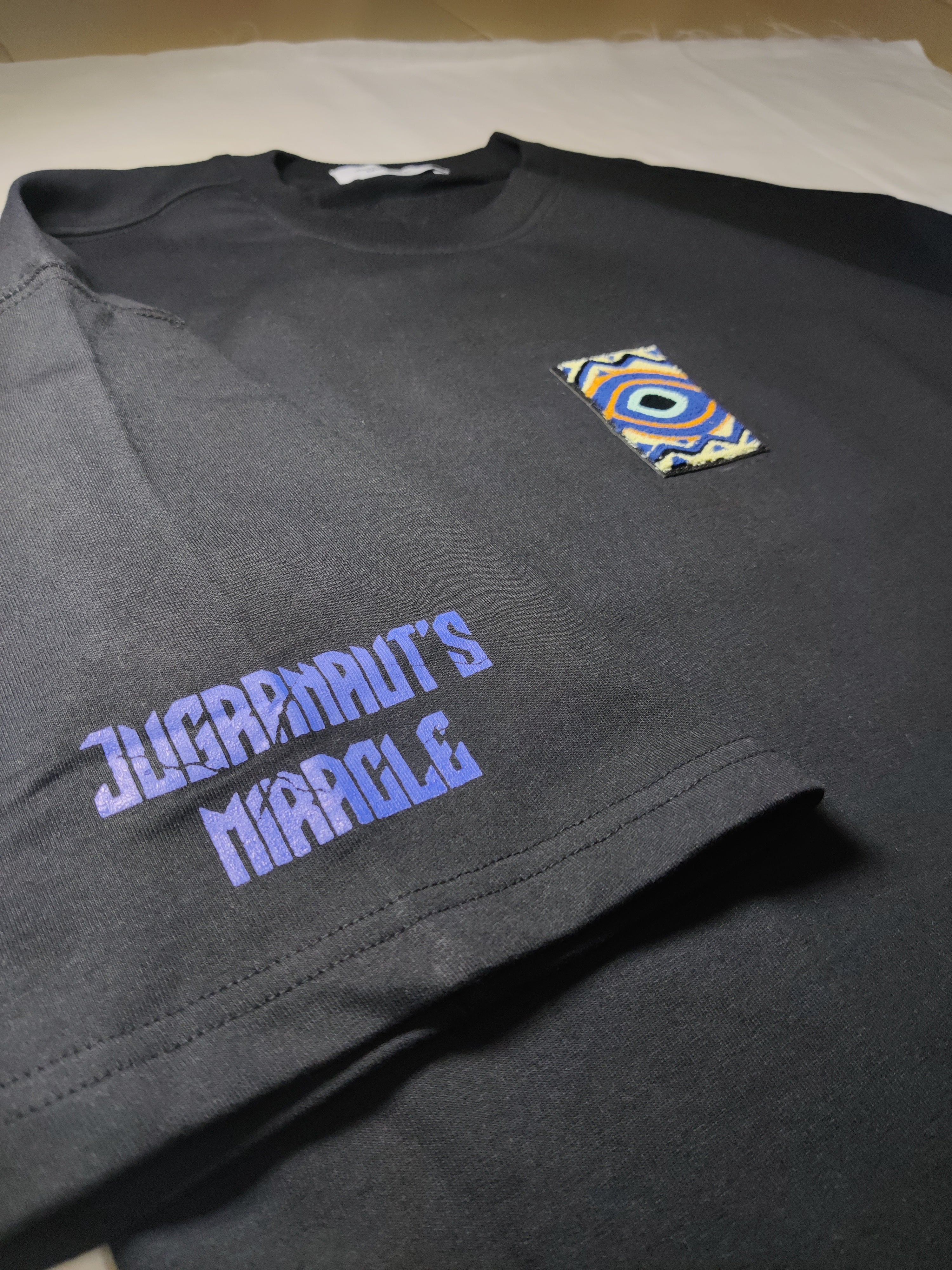 Dark gray long-sleeve shirt with 'Jugrnaut's Miracle' text and colorful patch on a white background