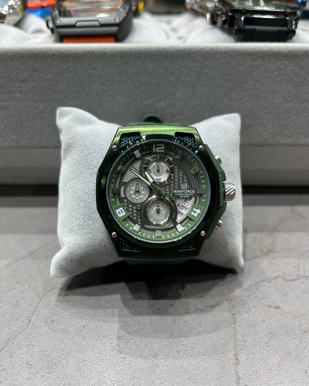Green watch on a gray cushion with a blurred background of other watches.