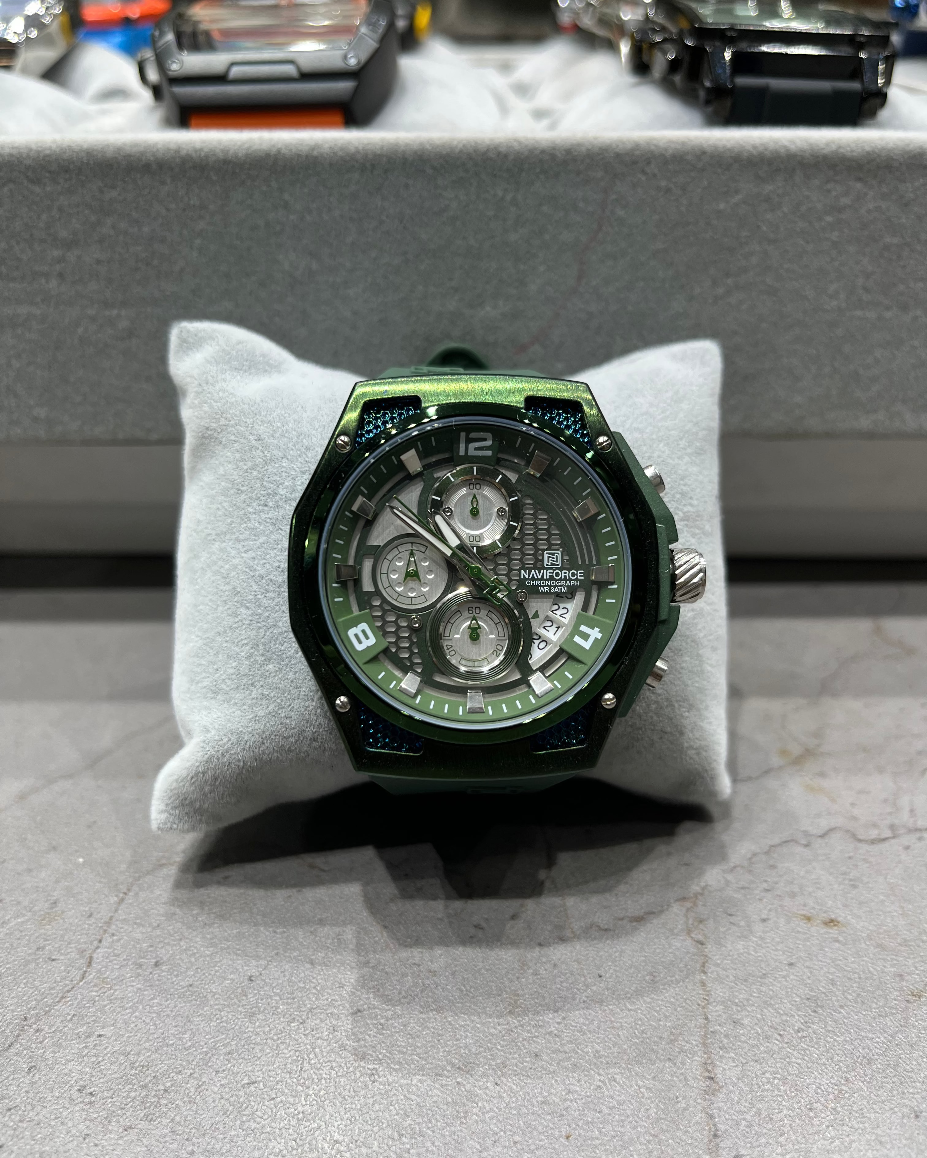 Green watch on a gray cushion with a blurred background of other watches.
