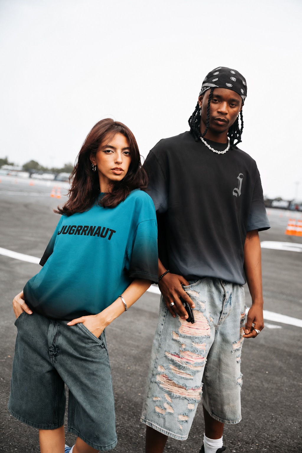 Two people standing on a road, one wearing a teal shirt with text and the other in a black shirt and ripped jeans.