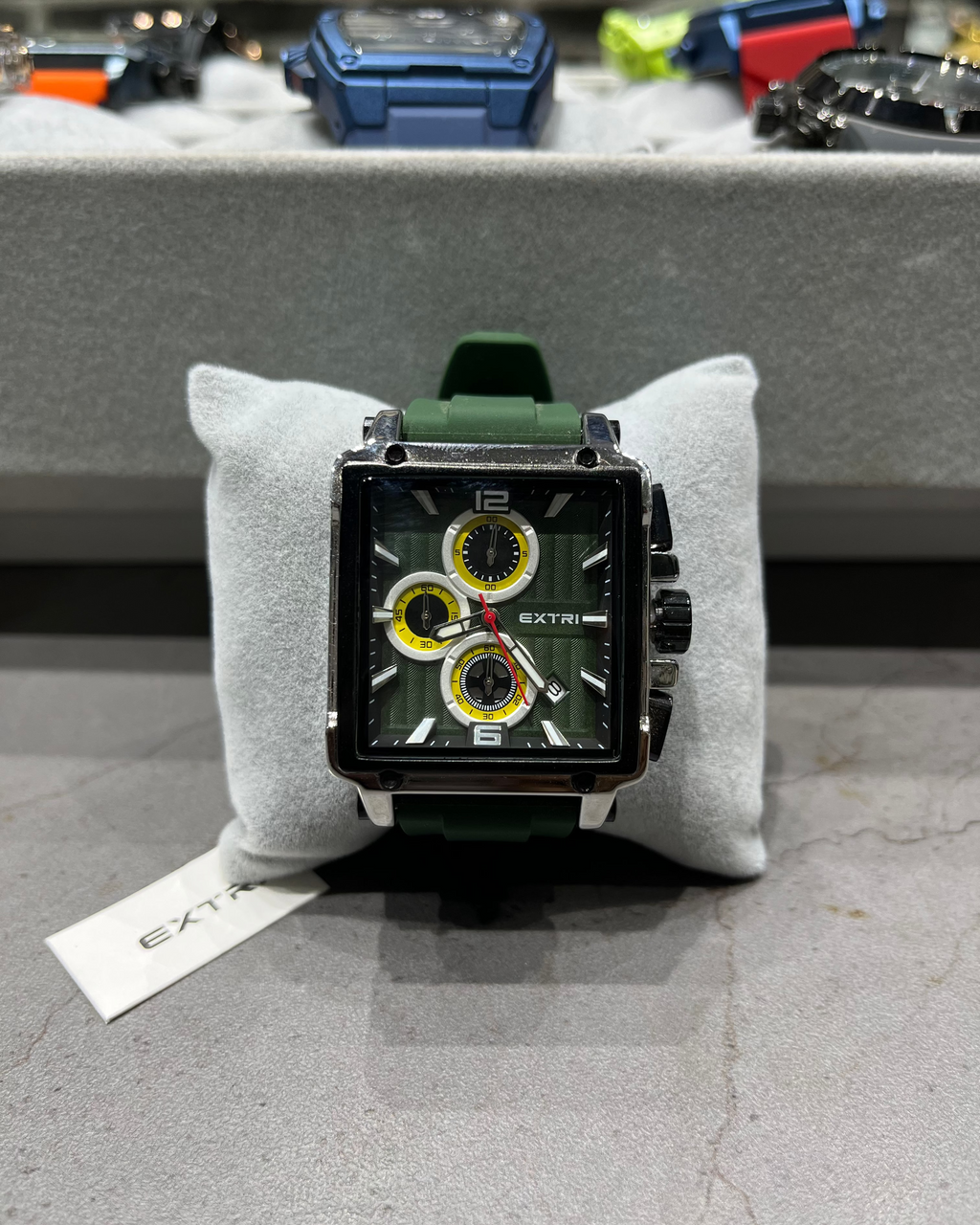 Watch with a green strap on a gray cushion, brand name visible, with a blurred background of other watches.