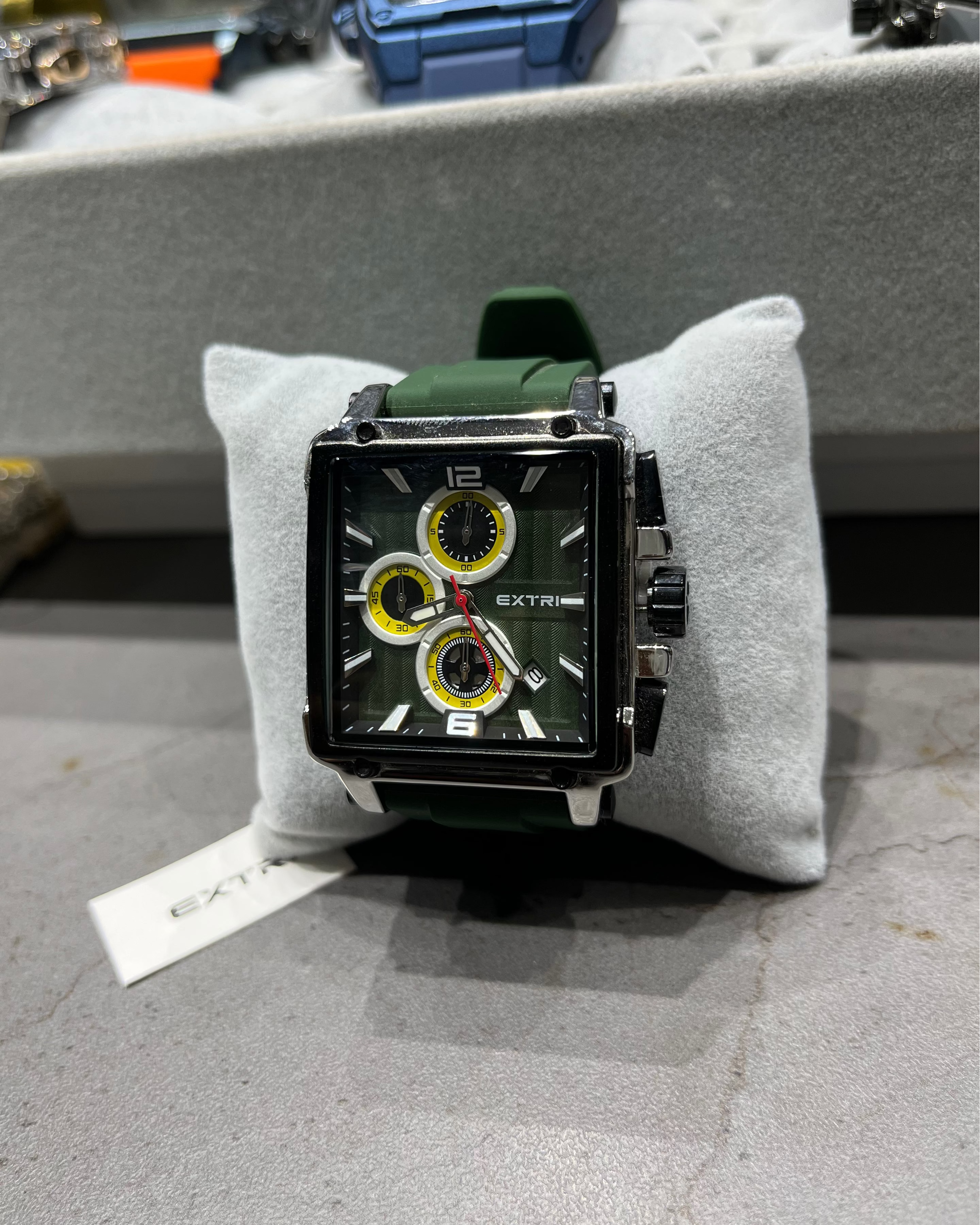 Watch with green strap on a gray cushion, with a blurred background of other watches.
