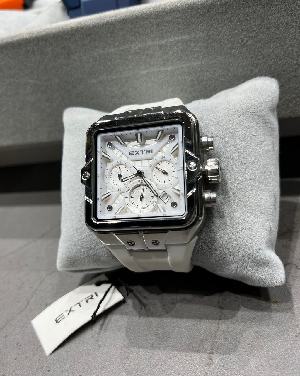 Silver watch with detailed face on a gray cushion, brand tag visible, on a gray surface.