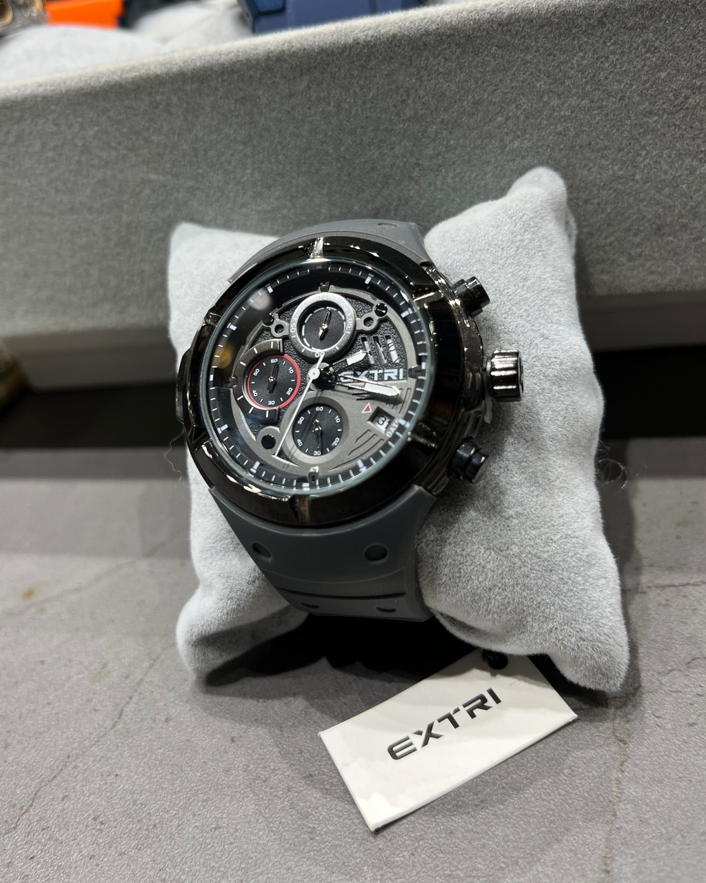 Luxury watch with visible brand name on a gray cushion, with a blurred background of other watches.