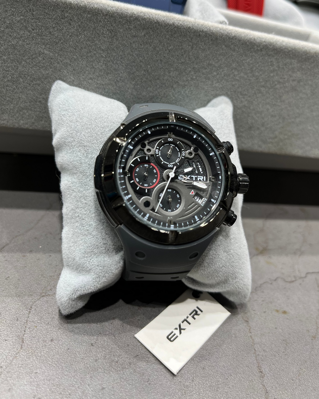 Black wristwatch on a gray cushion with 'EXTRI' branding, surrounded by other watches in the background.