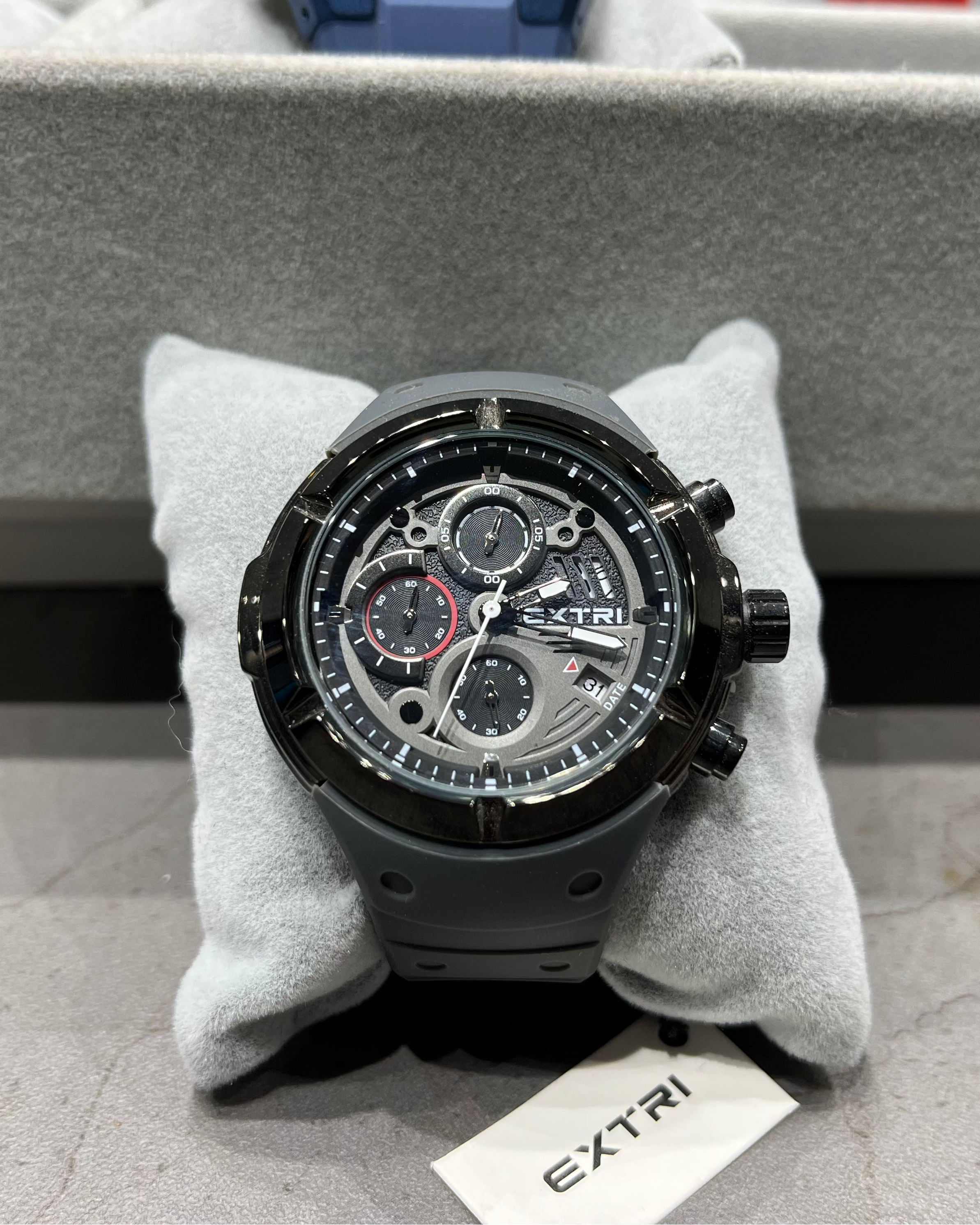 Black Extri watch on a gray cushion with a branded box in the background