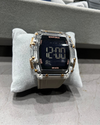 Digital watch with clear case and gold accents on a gray cushion, with colorful objects in the background.