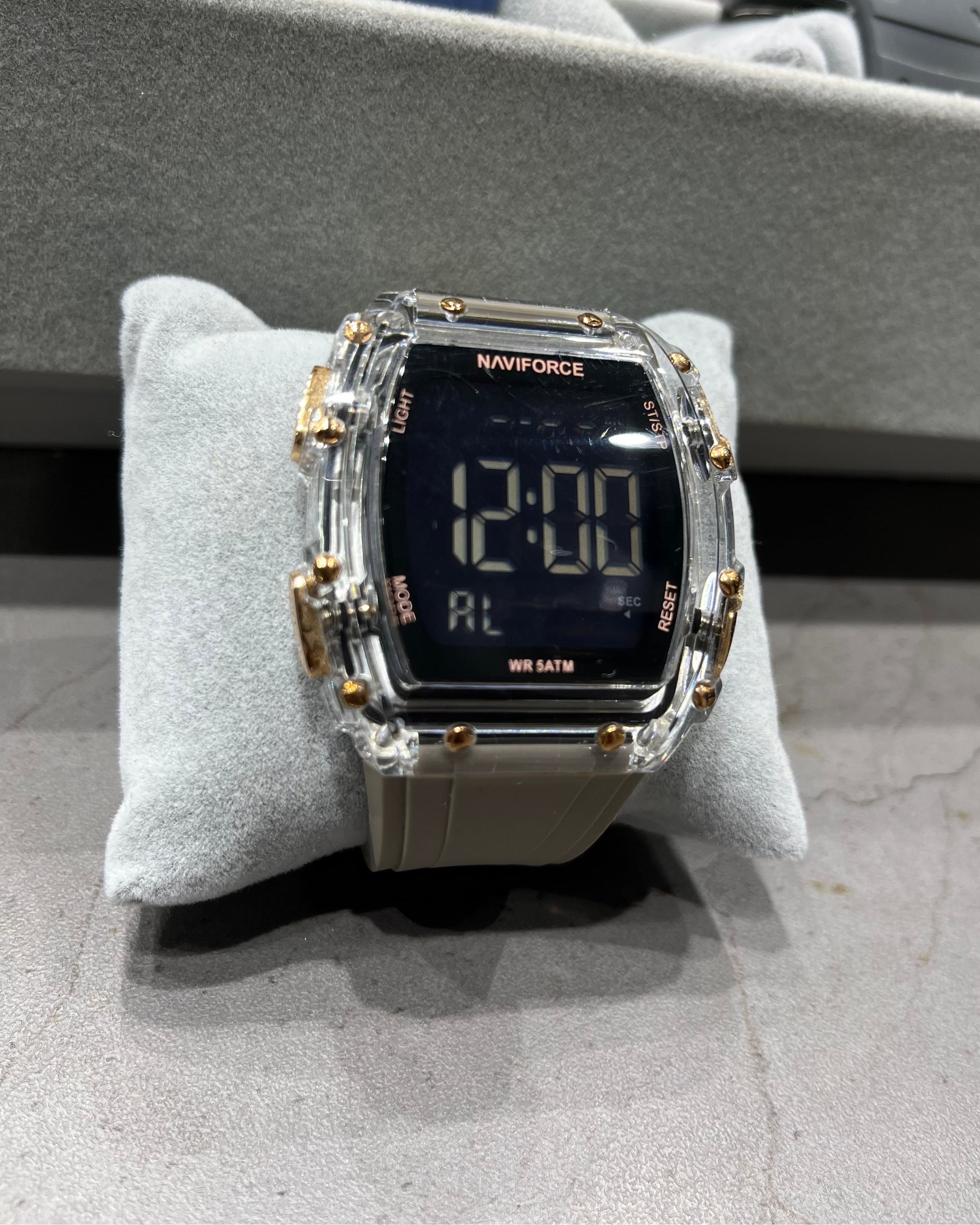 Digital watch with clear case and gold accents on a gray cushion, with colorful objects in the background.