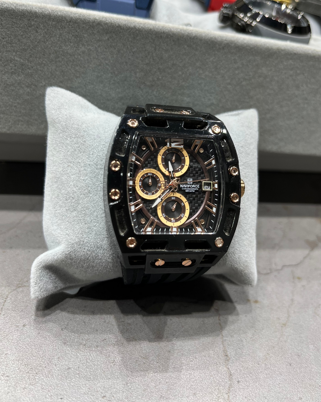 Luxury watch with black and gold design on a gray cushion, with other watches in the background.