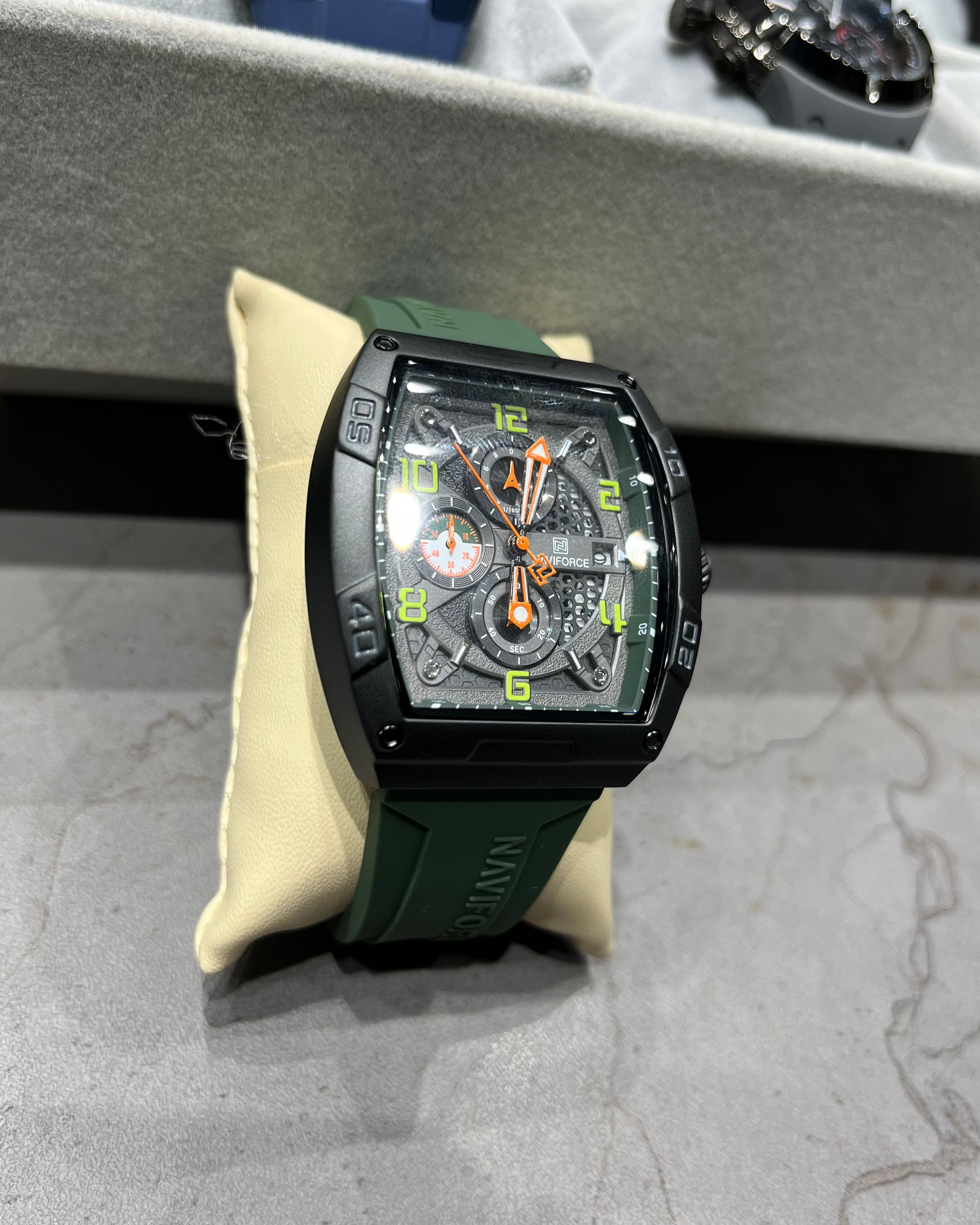 Black watch with green strap on a gray surface, with other watches in the background.