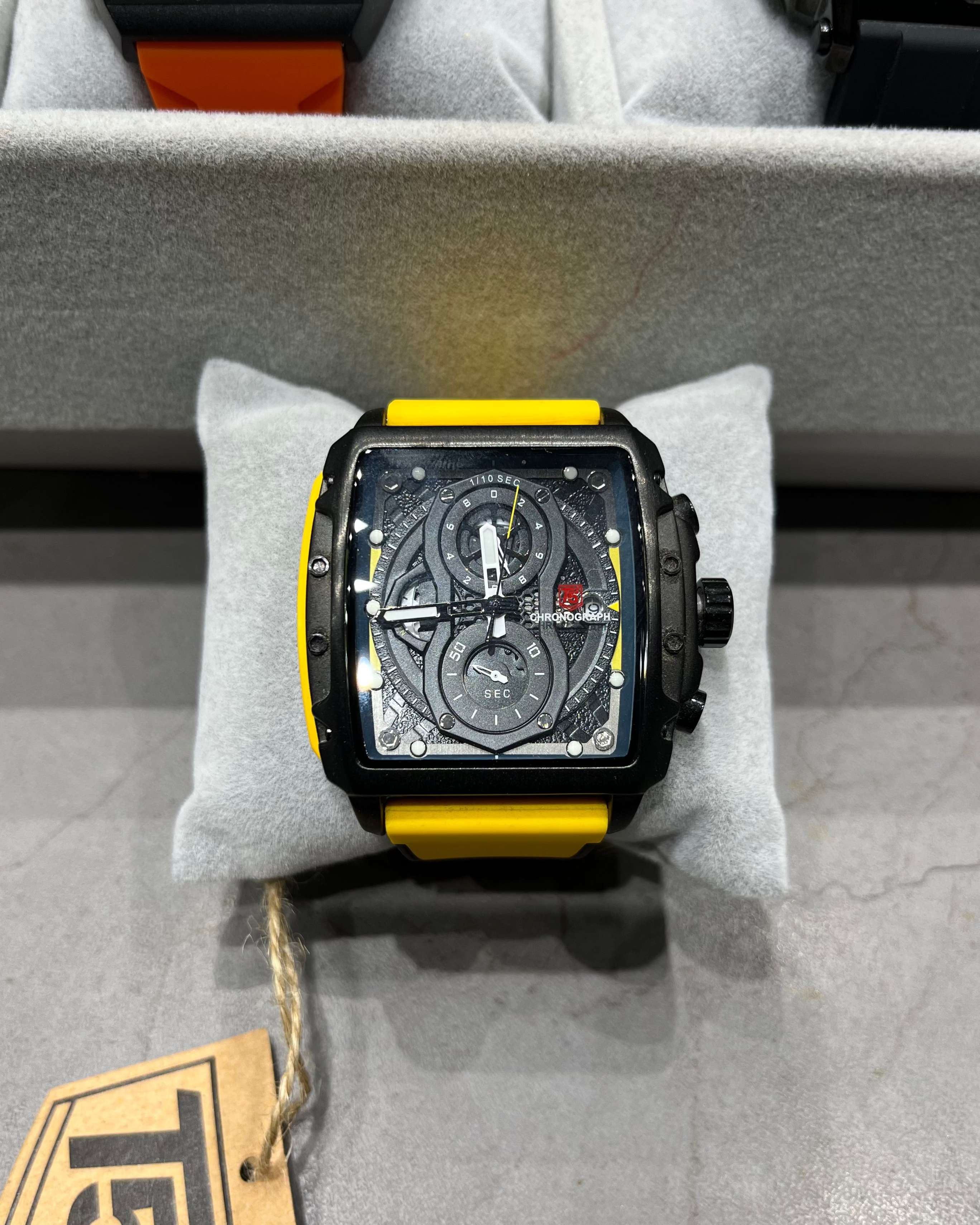 Luxury watch with black and yellow strap on a gray cushion, brand tag visible.