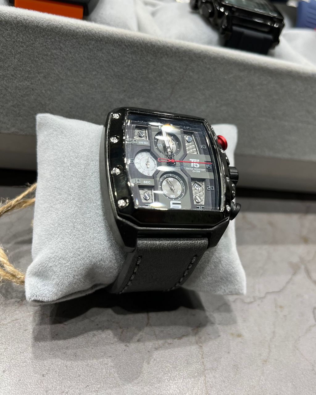 Black watch with detailed face on a gray cushion, with blurred background of other watches.