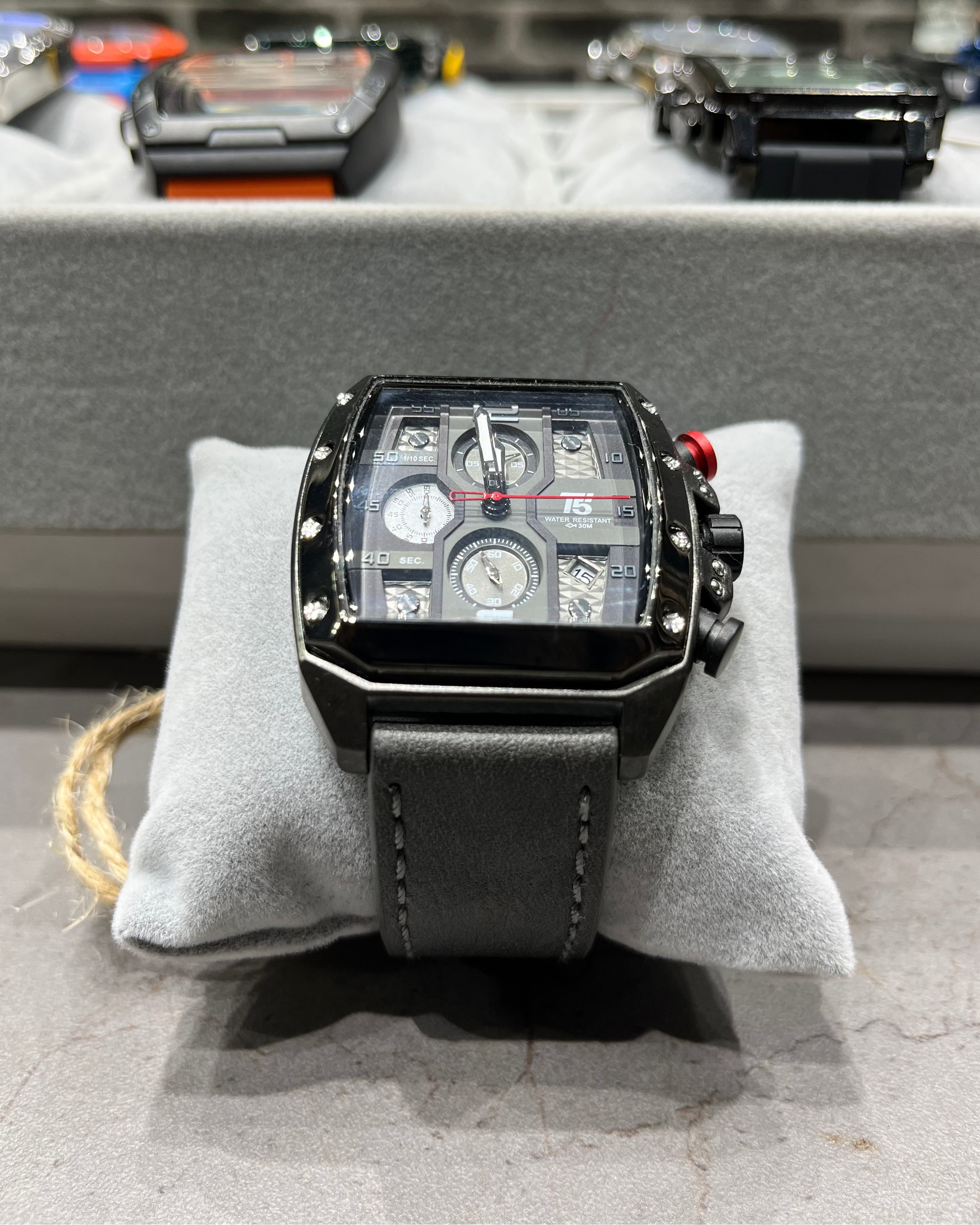 Luxury watch with black strap on a gray cushion, blurred background of other watches.